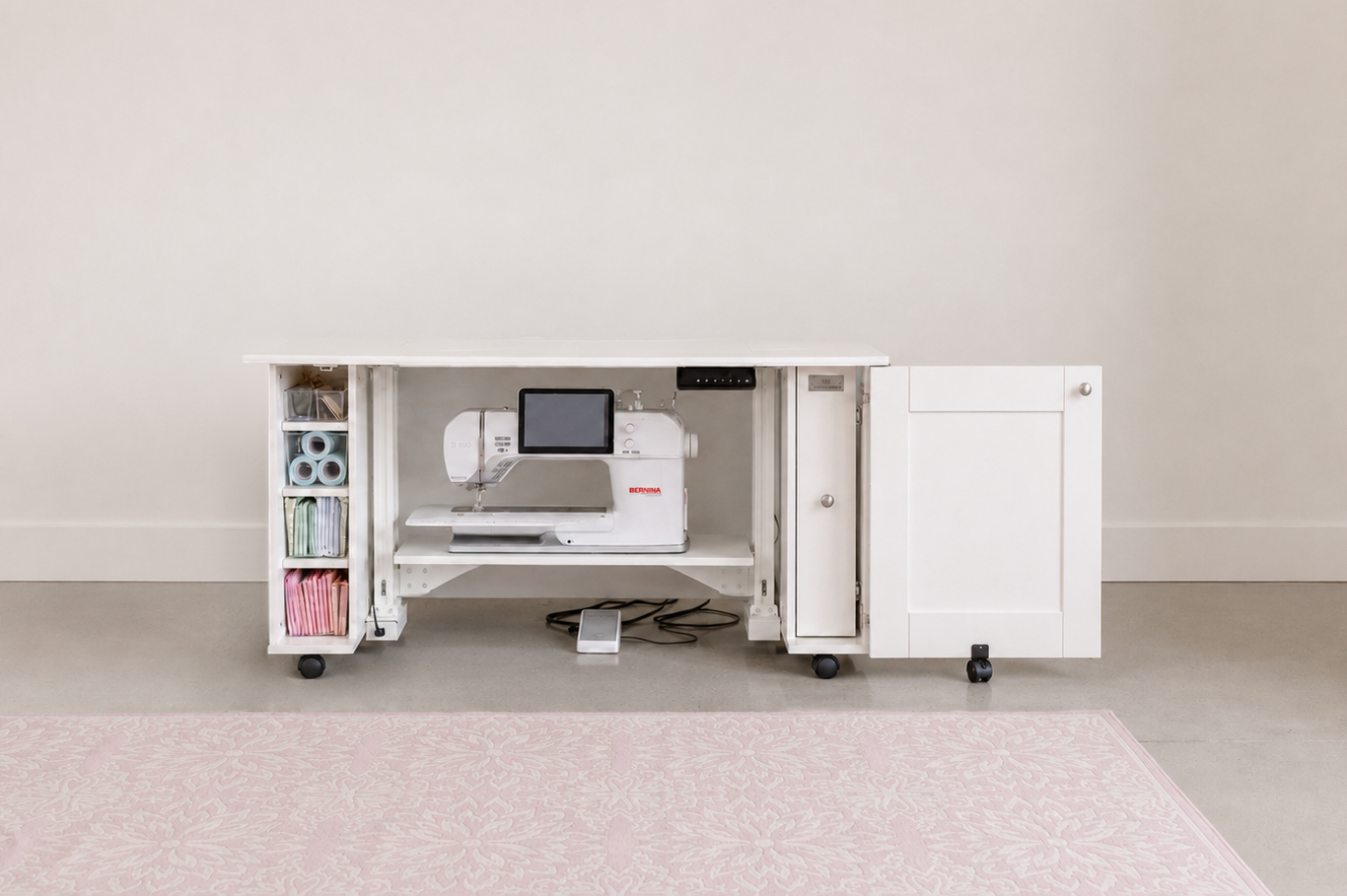 A white under-desk sewing machine cabinet with open shelves and a side cabinet door, shown in a bright room with a light pink rug. A white sewing machine is placed inside the desk. [xl]