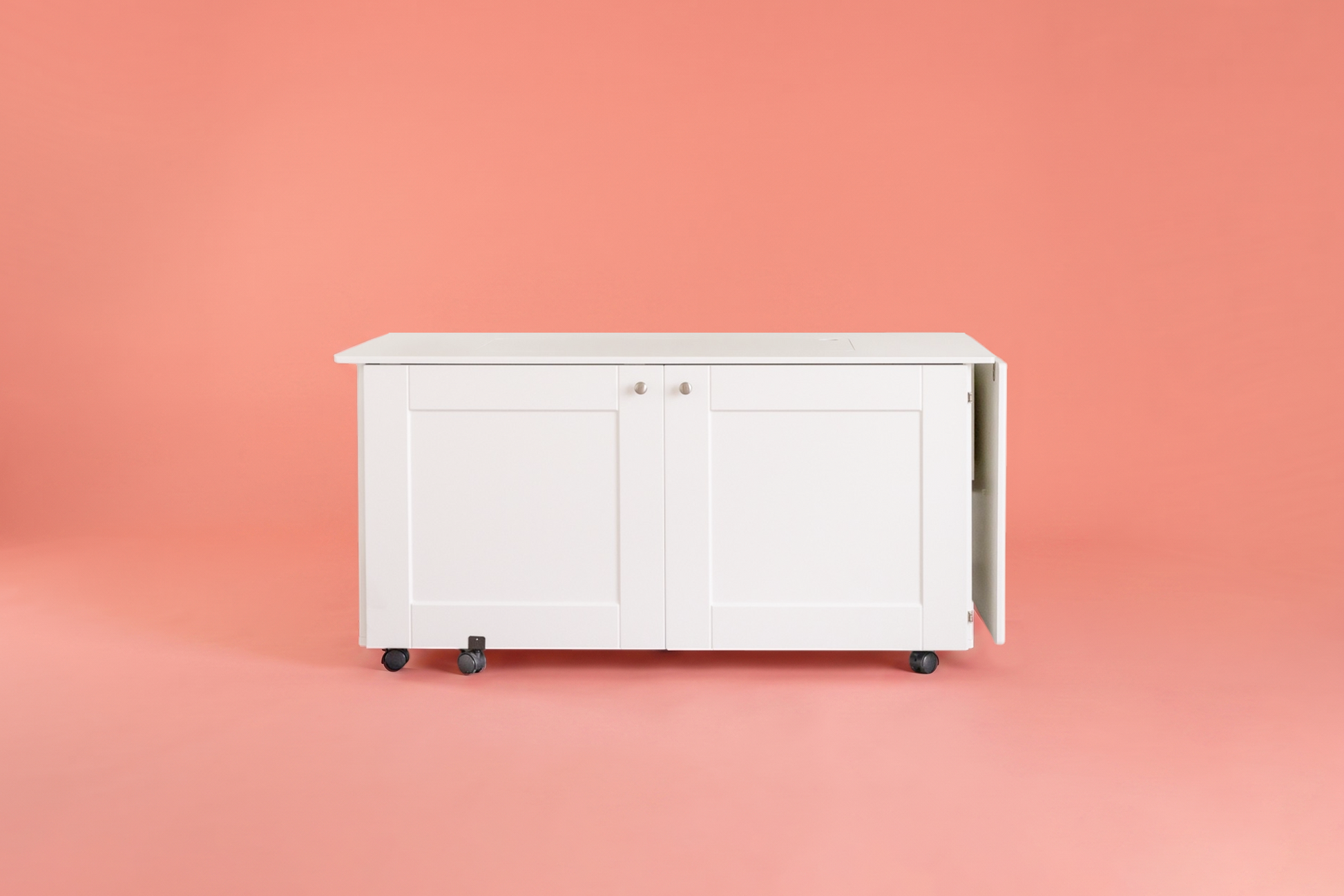 A white rolling storage cabinet with two paneled doors and black caster wheels, shown against a solid coral-pink background. [XL]